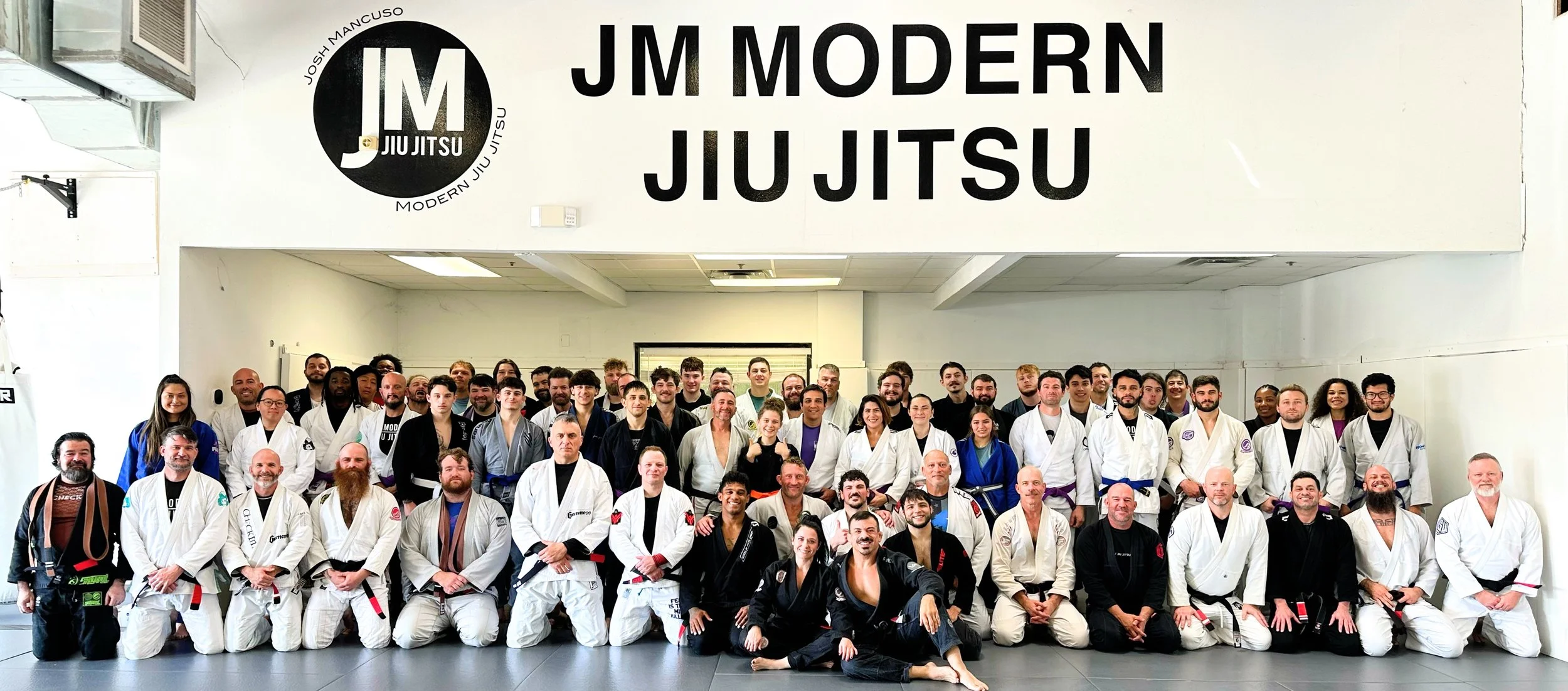 The adult class at JM Modern Jiu Jitsu gathered under the academy sign